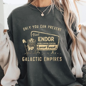 Puede incluir: Sudadera gris oscuro con un gráfico de estilo vintage. El diseño presenta el texto "ONLY YOU CAN PREVENT" encima de un letrero que dice "Visit ENDOR, Hiking-Camping-Feasting, Lunar Forest." Debajo está "GALACTIC EMPIRES."