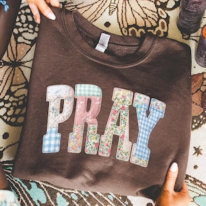 Op de afbeelding: Een bruine sweatshirt met het woord "PRAY" op de voorkant. De letters zijn gemaakt van patchwork stof met verschillende bloemen- en ruitpatronen. De sweatshirt wordt getoond.
