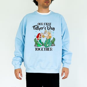 Puede incluir: Sudadera azul claro con un dise&ntilde;o de Ariel y el Rey Trit&oacute;n, con el texto "OUR FIRST Father's Day TOGETHER". El dise&ntilde;o incluye elementos en negro, verde, rojo y dorado.