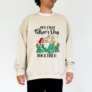 Puede incluir: Sudadera beige con un dise&ntilde;o de Ariel y el Rey Trit&oacute;n de La Sirenita, con el texto "OUR FIRST Father's Day TOGETHER". La ilustraci&oacute;n incluye algas verdes y burbujas azules.