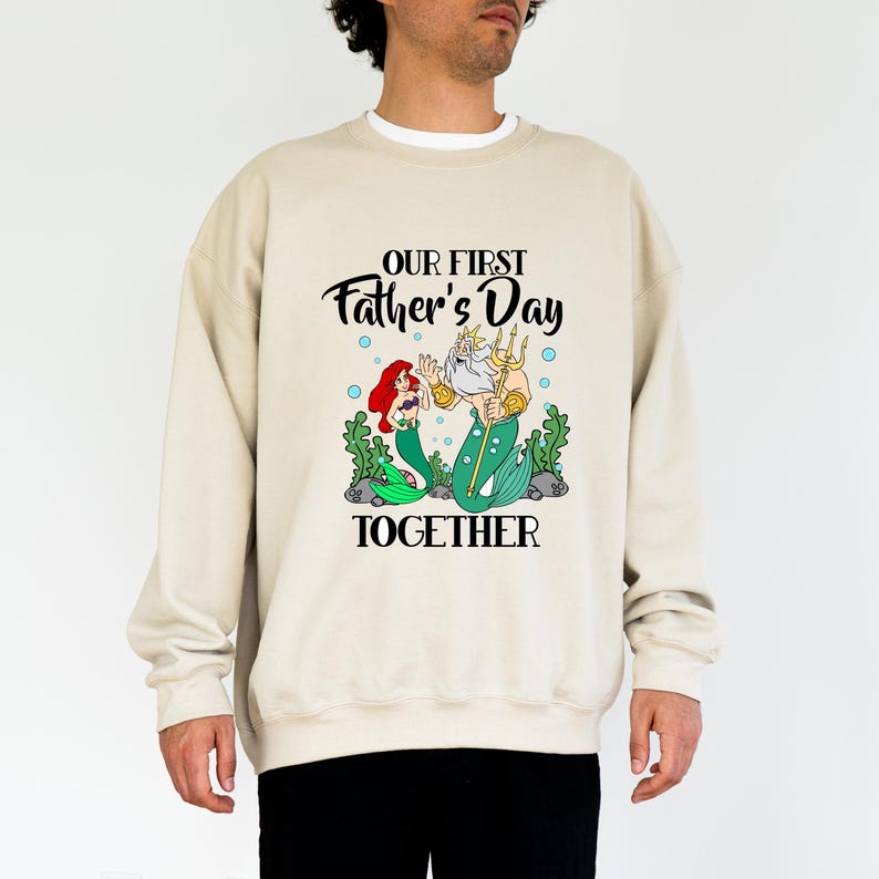 Puede incluir: Sudadera beige con un dise&ntilde;o de Ariel y el Rey Trit&oacute;n de La Sirenita, con el texto "OUR FIRST Father's Day TOGETHER". La ilustraci&oacute;n incluye algas verdes y burbujas azules.