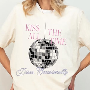 Op de afbeelding: Crèmekleurig T-shirt met een discobal-afbeelding. De tekst "KISS ALL THE TIME" staat in roze boven de discobal, met "Disco. Occasionally" in een cursief lettertype eronder. Het shirt heeft korte mouwen.