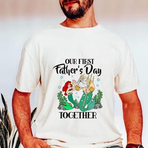 Puede incluir: Camiseta color crema con una ilustraci&oacute;n de dibujos animados de una sirena y el Rey Trit&oacute;n con el texto "OUR FIRST Father's Day TOGETHER". El gr&aacute;fico incluye algas verdes y burbujas azules.