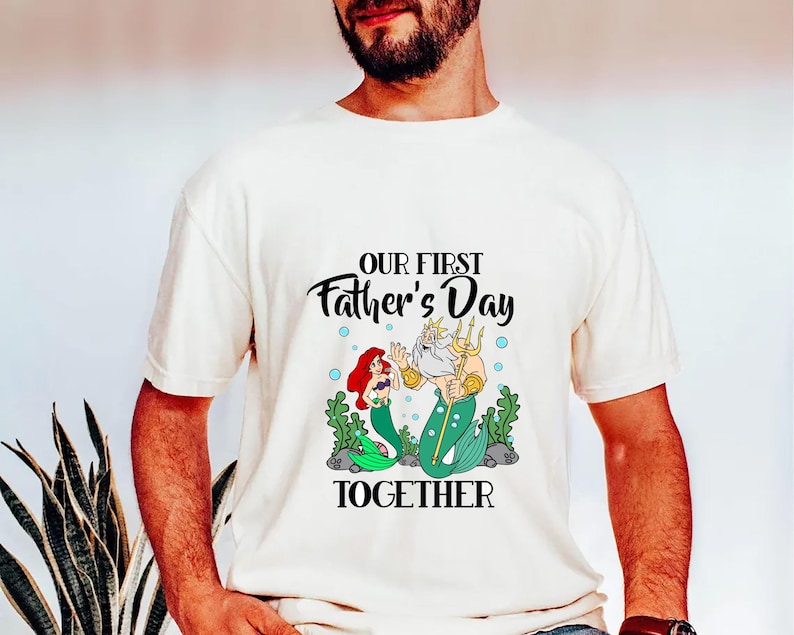 Puede incluir: Camiseta color crema con una ilustraci&oacute;n de dibujos animados de una sirena y el Rey Trit&oacute;n con el texto "OUR FIRST Father's Day TOGETHER". El gr&aacute;fico incluye algas verdes y burbujas azules.