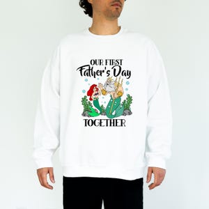 Puede incluir: Sudadera blanca con un gr&aacute;fico de Ariel y el Rey Trit&oacute;n de La Sirenita, con el texto "OUR FIRST Father's Day TOGETHER". El dise&ntilde;o incluye algas verdes y burbujas azules. Un art&iacute;culo de ropa con tem&aacute;tica del D&iacute;a del Padre.