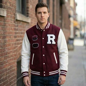 Op de afbeelding: Een bordeauxrood varsity-jack met witte mouwen, voorzien van een grote witte letter R op de borst, twee geborduurde patches, een drukknoopsluiting en gestreepte ribboorden bij de kraag, mouwen en zoom, gedragen over een donkerblauwe spijkerbroek.