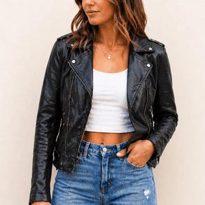 May include: A black faux leather motorcycle jacket with silver-tone metal zips and snap button lapels, worn over a white ribbed crop top and blue denim jeans with a distressed detail on the thigh area, featuring a classic collar and long sleeves.