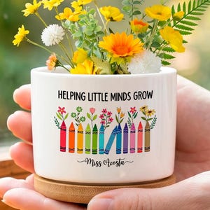May include: A white ceramic planter with a bamboo base containing a bouquet of yellow, orange, and white flowers. The planter features the text "HELPING LITTLE MINDS GROW" above a graphic of colorful crayons topped with various small floral illustrations.