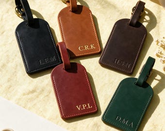 Puede incluir: Cinco etiquetas de equipaje rectangulares de cuero en negro, tostado, marrón oscuro, granate y verde oscuro. Cada etiqueta tiene una parte superior redondeada, una correa de cuero a juego con un anillo de metal color latón e iniciales doradas: L.E.M, C.R.K, H.S.N, V.P.L y D.M.A.