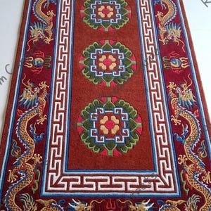 Könnte beinhalten: Ein rechteckiger roter Teppich mit detailliertem Design. Der Teppich hat eine zentrale Fläche mit drei Blumenmustern und einen Rand mit Drachen und geometrischen Mustern. Die Drachen sind gold, blau und grün.
