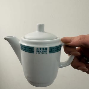 Pode incluir: Bule de cerâmica branco com tampa e alça curva. O bule tem uma faixa turquesa e branca com o texto "PEKING RESTORAN" em inglês e caracteres chineses.