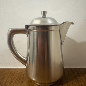 May include: A brushed stainless steel coffee pot with a curved handle, a lid, and a spout. The pot has a cylindrical body and sits on a wooden surface. The handle is arched and the lid has a small knob.