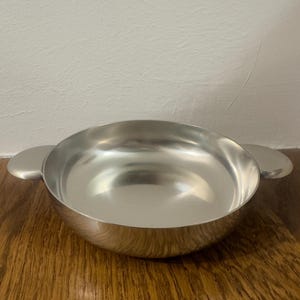 Może przedstawiać: Błyszcząca, srebrna miska z dwoma małymi, zaokrąglonymi uchwytami po bokach. Miska ma gładką, odbijającą światło powierzchnię i stoi na drewnianej powierzchni. Projekt jest prosty i nowoczesny, odpowiedni do serwowania lub ekspozycji. Średnica wynosi około 15 cm.