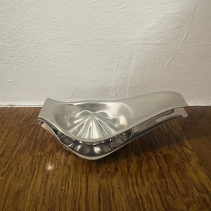 Op de afbeelding: Een zilverkleurige citruspers met een geribbelde, kegelvormige ruimer in het midden. De pers heeft een gebogen ontwerp met een tuit voor gemakkelijk schenken. Het product is gemaakt van metaal en staat op een houten oppervlak.