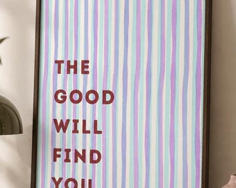 Puede incluir: Un cuadro rectangular con marco que presenta rayas verticales en tonos alternos de violeta claro y azul claro. El texto "THE GOOD WILL FIND YOU" está impreso en una fuente sans-serif negrita de color rojo oscuro sobre el fondo a rayas.