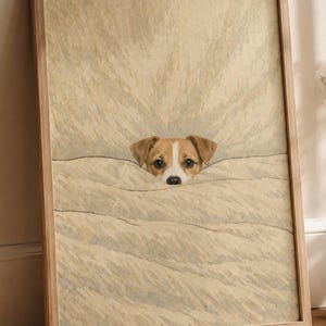 Puede incluir: Una impresión enmarcada que presenta la cabeza de un perro asomando por debajo de una manta. El perro tiene pelaje marrón y blanco, ojos oscuros y una nariz negra. La manta es de color beige claro y el marco es de madera clara.