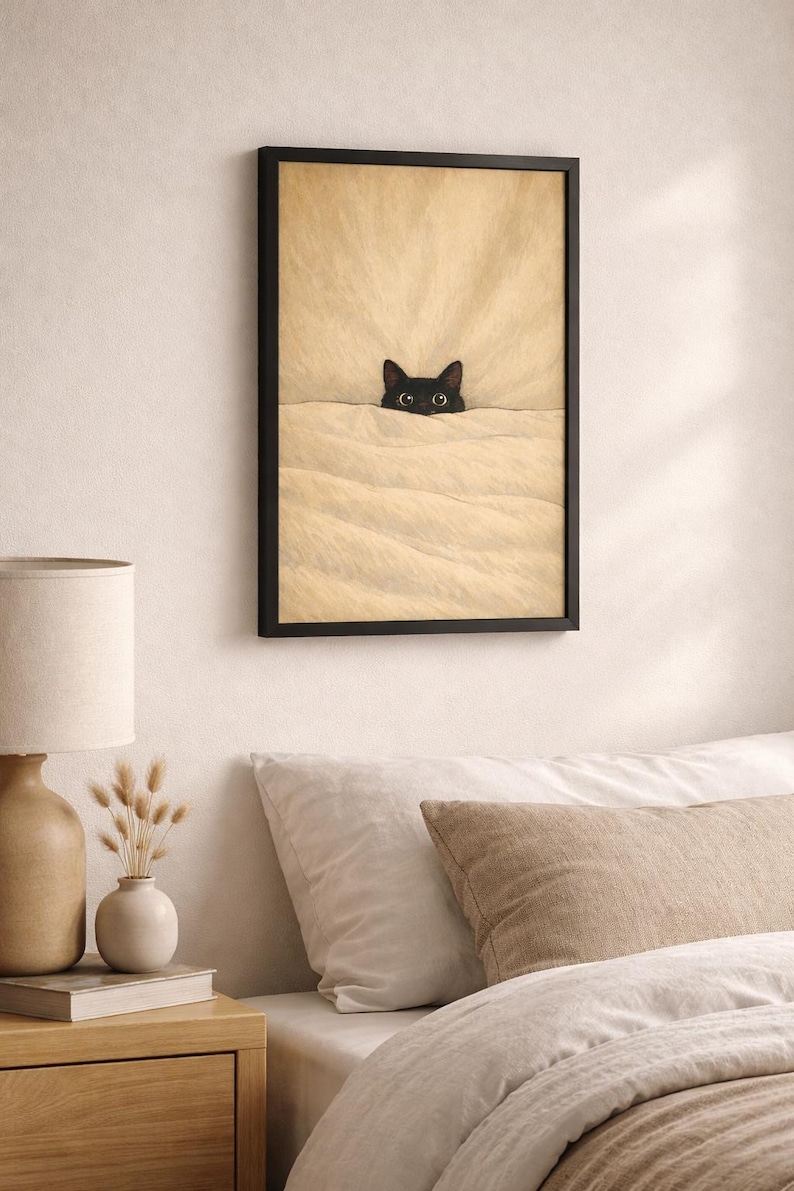Puede incluir: L&aacute;mina enmarcada que muestra un gato negro asomando por encima de una manta beige. Se ven los ojos del gato, a&ntilde;adiendo un toque de fantas&iacute;a. La obra est&aacute; enmarcada en negro, ideal para la decoraci&oacute;n de dormitorios o salones.