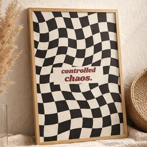 Puede incluir: Una impresión artística enmarcada con un patrón de cuadros en blanco y negro que parece ondulado. Las palabras "controlled chaos." están impresas en color burdeos en el centro. El marco es de color marrón claro.
