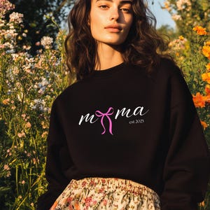 Może przedstawiać: Czarna bluza z napisem "mama" w ozdobnym kroju pisma i różową kokardą. Tekst "est 2025" znajduje się pod słowem "mama". Modelka ma na sobie kwiecistą spódnicę i jest otoczona kwiatami.