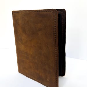 May include: A dark brown leather cover for an electronic device, featuring a matte, textured surface with visible stitching along the edges. The cover is shown in a partially open position, revealing a dark interior frame that holds the device securely.