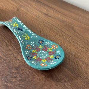 Może przedstawiać: Turkusowa ceramiczna podstawka pod łyżkę z dekoracyjnym wzorem kwiatowym w kolorze czerwonym, żółtym, białym i niebieskim. Podstawka pod łyżkę ma zakrzywiony uchwyt i szeroką, owalną miseczkę. Przedmiot umieszczono na drewnianej powierzchni.