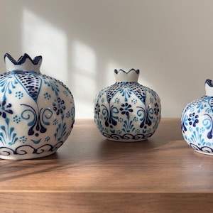 Peut inclure: Trois objets décoratifs en céramique en forme de grenade. Les objets blancs présentent des motifs floraux bleus et un sommet en forme de couronne. Ils sont disposés sur une surface en bois, avec un vase de fleurs séchées en arrière-plan.