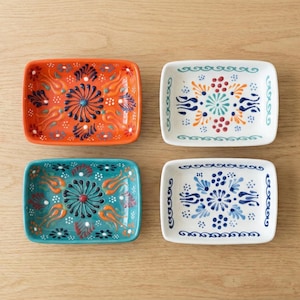 Könnte beinhalten: Vier rechteckige Keramikschalen mit floralen Mustern. Die Schalen sind orange, türkis und weiß. Die orangefarbene Schale hat ein blaues und weißes Blumenmuster. Die türkisfarbene Schale hat ein rot-weiß-blaues Blumenmuster. Die weißen Schalen haben blau-orangefarbene Blumenmuster.