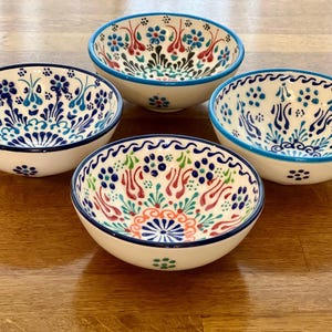 Może przedstawiać: Cztery małe, okrągłe ceramiczne miski ze skomplikowanymi wzorami kwiatowymi w kolorze niebieskim, czerwonym i zielonym na białym tle. Każda miska ma ciemnoniebieską lub turkusową obwódkę. Miski są ułożone na drewnianej powierzchni.