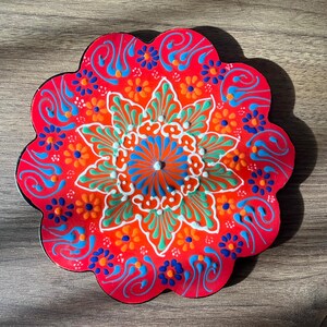 Puede incluir: Un plato decorativo circular con bordes festoneados y una base de color rojo vibrante. La superficie está adornada con un patrón de mandala floral simétrico y multicolor en tonos de pintura naranja, azul claro, azul oscuro, blanco y verde.
