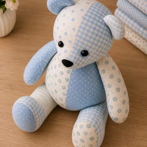 May include: A stuffed teddy bear crafted from various light blue and white fabrics featuring patterns of small stars, gingham checks, polka dots, and floral motifs. The bear has black button eyes and a black nose, with a patchwork design throughout.