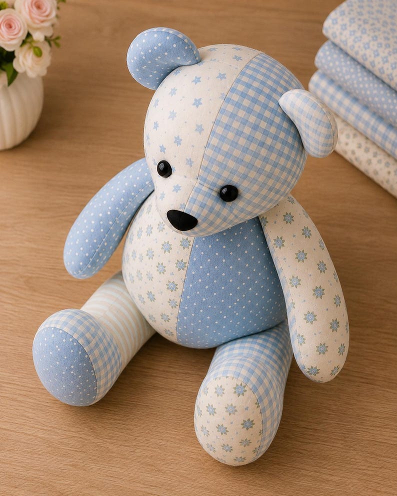 May include: A stuffed teddy bear crafted from various light blue and white fabrics featuring patterns of small stars, gingham checks, polka dots, and floral motifs. The bear has black button eyes and a black nose, with a patchwork design throughout.