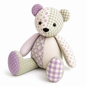 May include: A stuffed teddy bear constructed from various fabric patches in shades of lavender, sage green, and white. The bear features a mix of floral, polka dot, striped, and gingham patterns, with black plastic eyes and a black fabric nose.