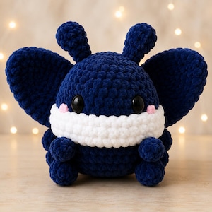 May include: A dark blue crocheted plush toy shaped like a moth or butterfly with large wings, two antennae, and a wide white band across the face. It features two large black plastic eyes and small pink stitched accents on the cheeks.