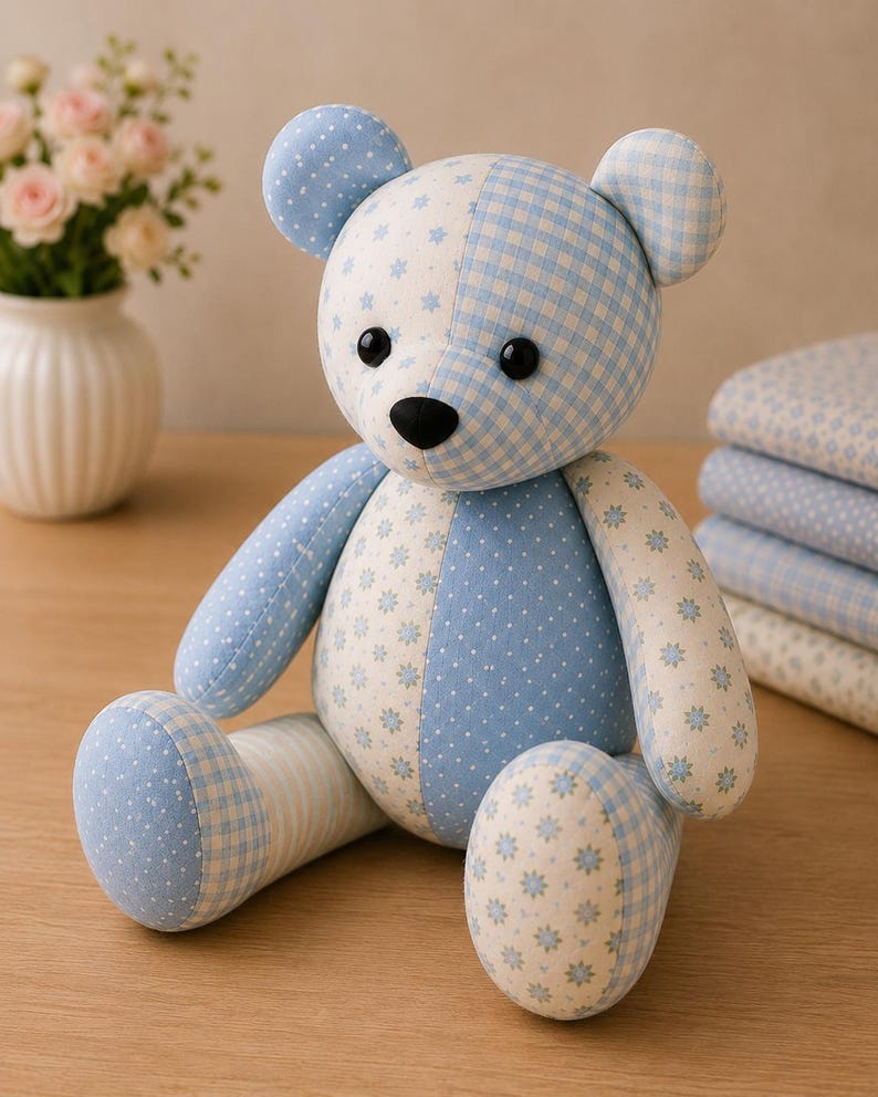 May include: A stuffed teddy bear crafted from various light blue and white fabrics featuring patterns of small flowers, polka dots, and gingham checks. The bear has two round black eyes and a black nose, with a seated posture and stitched seams.