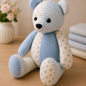 May include: A stuffed teddy bear constructed from various light blue and white fabric patches. The bear features a mix of patterns including gingham checks, small stars, and floral prints. It has two round black eyes and a black nose.