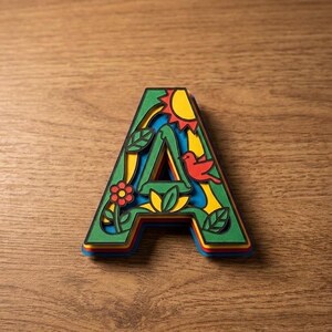 May include: A layered wooden letter "A" with a nature-themed design. The letter features a red bird, a yellow sun, and a flower. The edges are green, blue, yellow, and red, set against a wooden background.