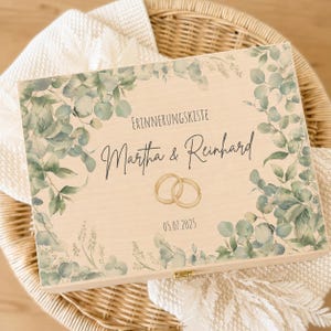 May include: A rectangular wooden keepsake box featuring a printed design of green eucalyptus leaves. The text reads "ERINNERUNGSKISTE", "Martha & Reinhard", and "05.07.2025". Two interlocking gold rings are illustrated in the center of the lid.