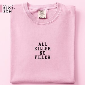 May include: A light pink t-shirt with the text "ALL KILLER NO FILLER" in black block letters. The shirt is folded neatly and has a Comfort Colors tag. The upper left corner has the text "COLOR BLOSSOM".