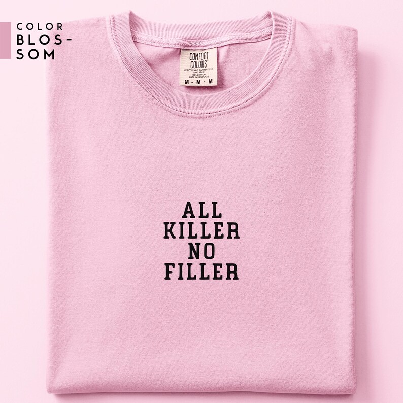 May include: A light pink t-shirt with the text "ALL KILLER NO FILLER" in black block letters. The shirt is folded neatly and has a Comfort Colors tag. The upper left corner has the text "COLOR BLOSSOM".