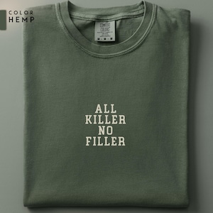 May include: A folded, olive green t-shirt with the text "ALL KILLER NO FILLER" in white block letters. The shirt has a crew neck and short sleeves. The brand tag is visible at the neckline.