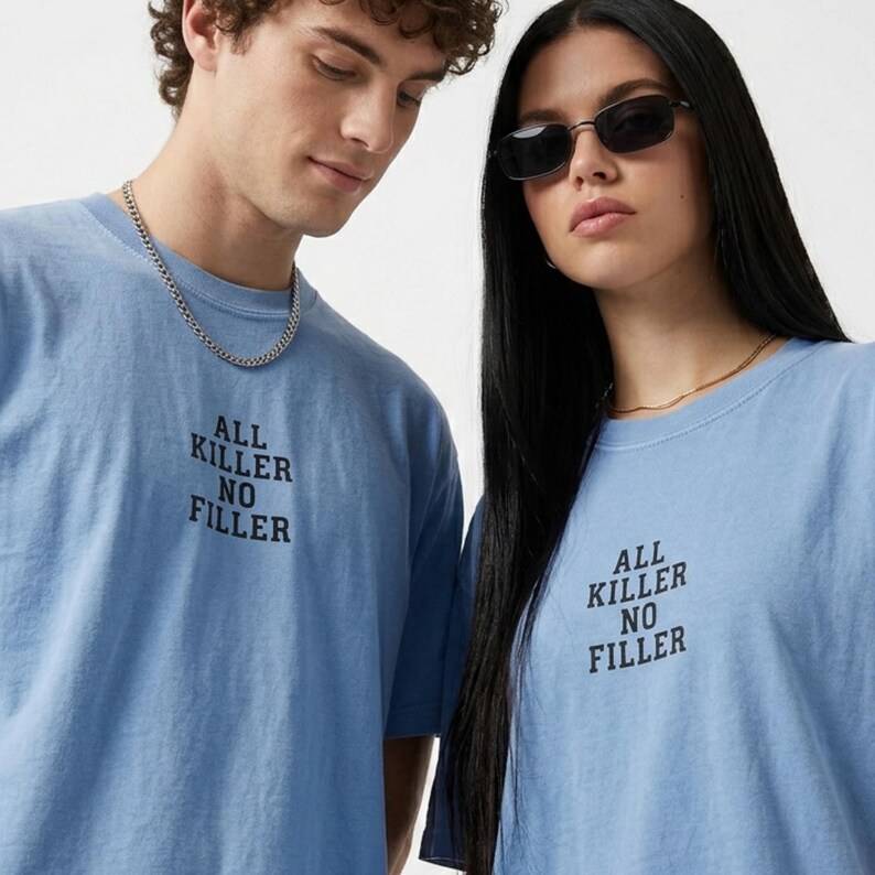 May include: Two people wearing light blue t-shirts with the text "ALL KILLER NO FILLER" in black. The shirts have short sleeves and a crew neck. One person is wearing a silver chain necklace, and the other is wearing sunglasses.