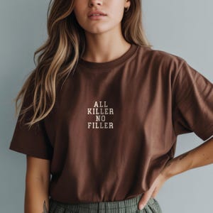 May include: A brown t-shirt with the text "ALL KILLER NO FILLER" in a white, sans-serif font. The shirt is a solid color and has a crew neck. The model is wearing the shirt.