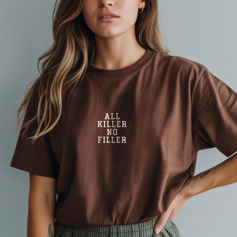 May include: A brown t-shirt with the text "ALL KILLER NO FILLER" in a white, sans-serif font. The shirt is a solid color and has a crew neck. The model is wearing the shirt.