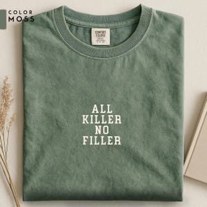 May include: A moss green t-shirt with the text "ALL KILLER NO FILLER" in white block letters. The shirt is folded neatly, with a small tag visible near the collar. The color swatch "COLOR MOSS" is in the upper left corner.