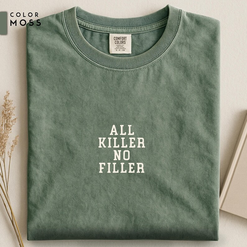 May include: A moss green t-shirt with the text "ALL KILLER NO FILLER" in white block letters. The shirt is folded neatly, with a small tag visible near the collar. The color swatch "COLOR MOSS" is in the upper left corner.