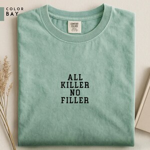 May include: A light green t-shirt with the text "ALL KILLER NO FILLER" in black block letters. The shirt has a crew neck and a small tag that says "COMFORT COLORS". The shirt is folded neatly on a light-colored surface.