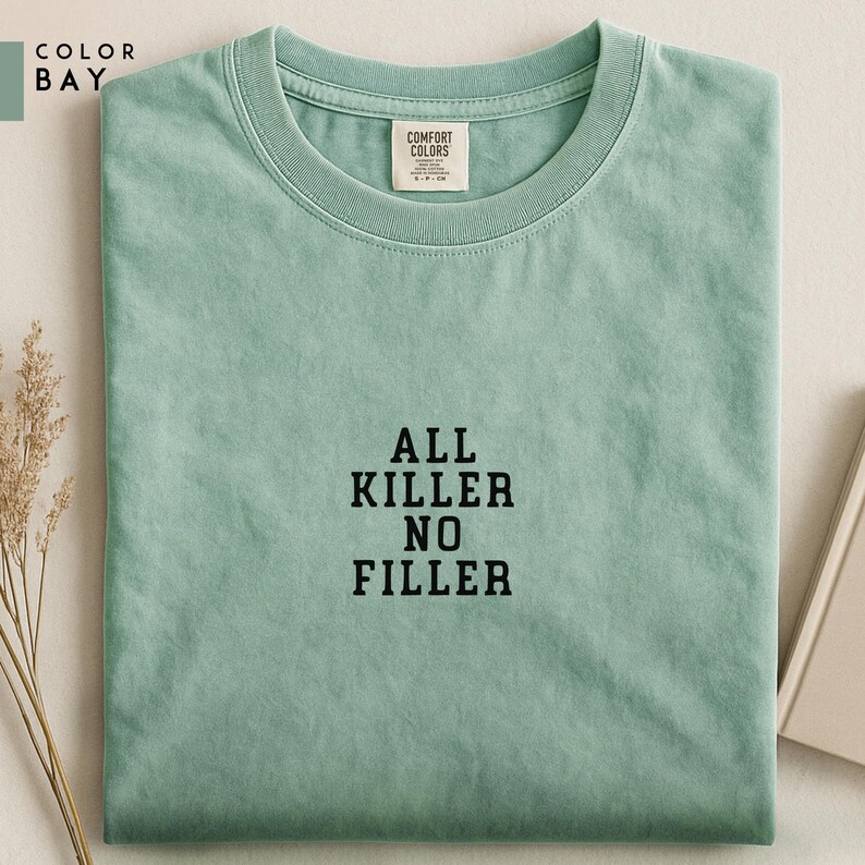 May include: A light green t-shirt with the text "ALL KILLER NO FILLER" in black block letters. The shirt has a crew neck and a small tag that says "COMFORT COLORS". The shirt is folded neatly on a light-colored surface.