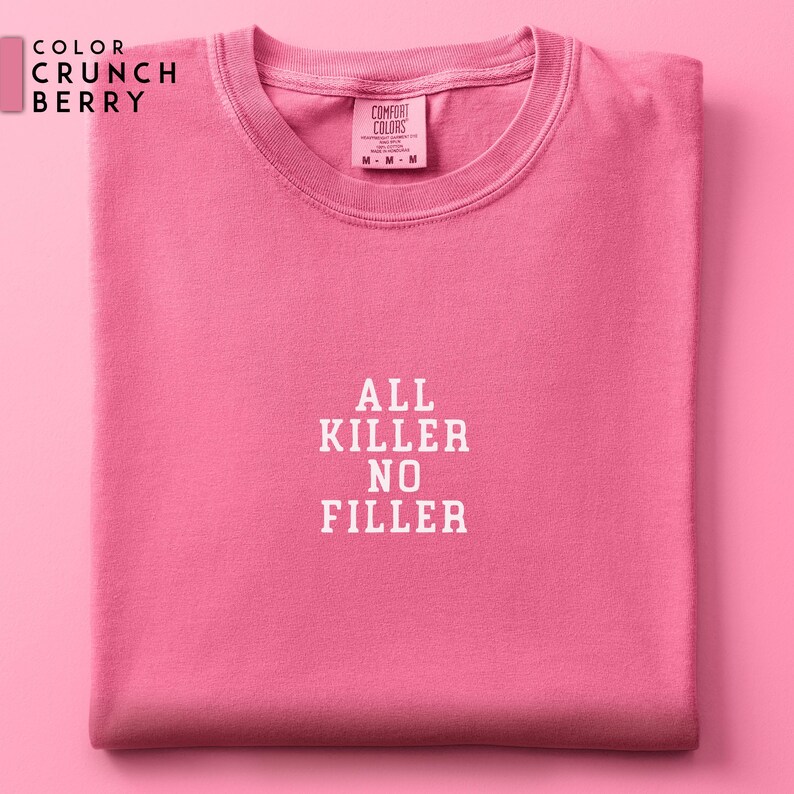 May include: A folded, short-sleeved, crew neck t-shirt in a vibrant Crunch Berry pink. The shirt features the text "ALL KILLER NO FILLER" in white block letters. The Comfort Colors brand tag is visible at the neckline.