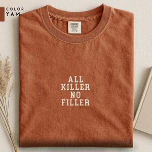 May include: A folded, rust-colored t-shirt with the text "ALL KILLER NO FILLER" in white block letters. The shirt has a crew neck and a small tag at the neckline. The color is labeled "YAM".
