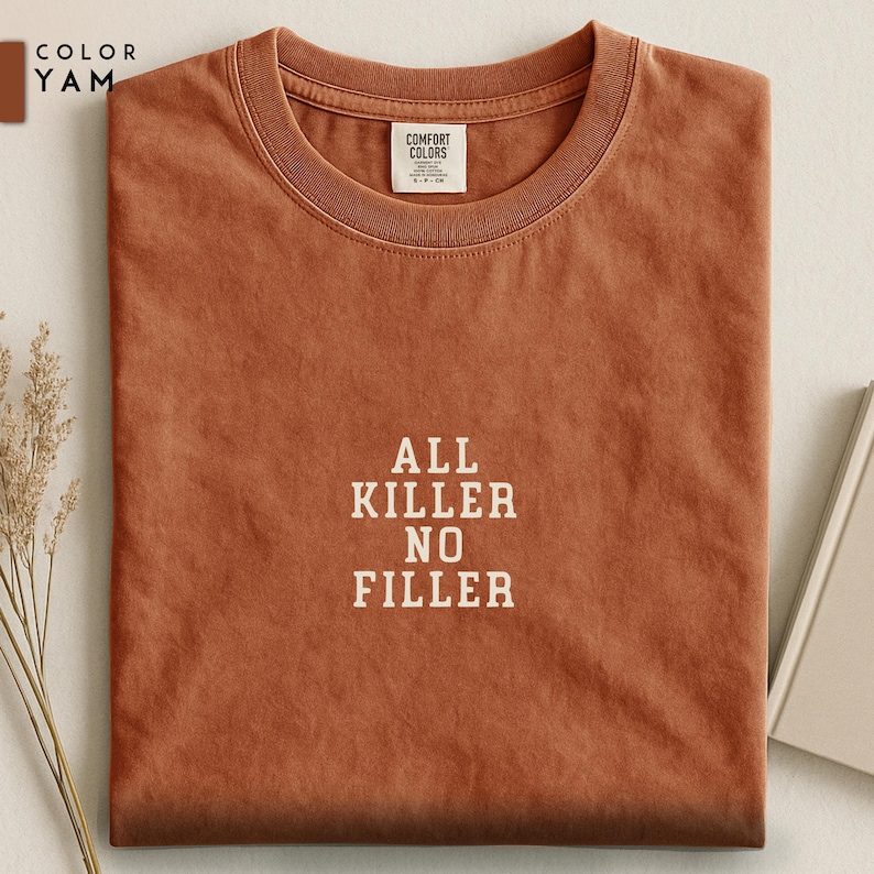 May include: A folded, rust-colored t-shirt with the text "ALL KILLER NO FILLER" in white block letters. The shirt has a crew neck and a small tag at the neckline. The color is labeled "YAM".